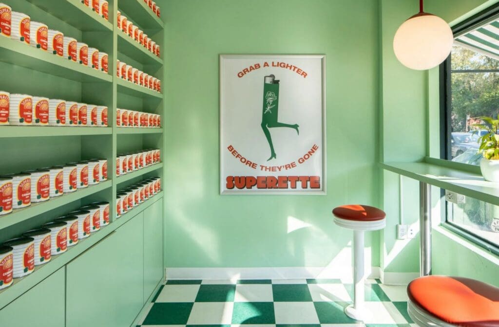Retro cannabis dispensary retail display with green shelves, checkerboard floor, branded product tins, and Superette wall art in a bright mint green storefront.