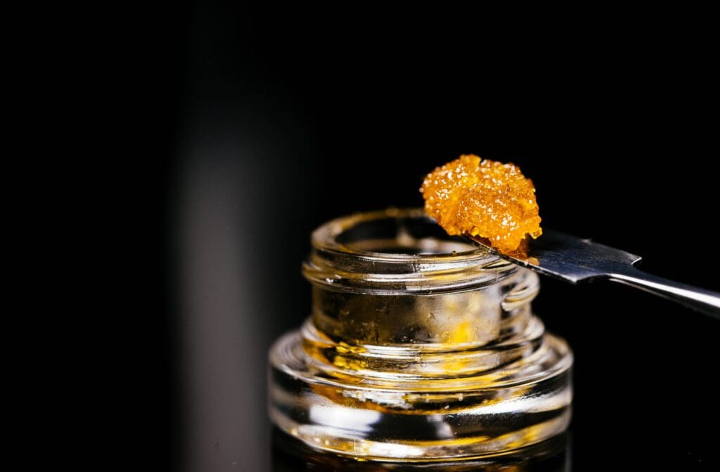 Cannabis concentrate in a glass jar with golden live resin on a dab tool against a dark background, highlighting the texture and quality of cannabis extracts for a blog about concentrates, dispensary products, cannabis retail, or industry trends.