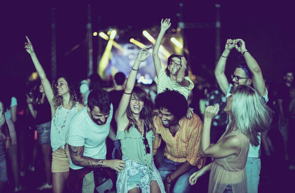 Group of friends smiling and dancing at an outdoor nighttime event with colorful stage lights in the background.